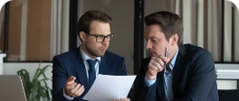 Dois homens discutindo algum acordo, um deles está segurando papel e o outro caneta, os dois estão sentados um de lao para o outro