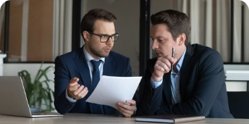 Dois homens discutindo algum acordo, um deles está segurando papel e o outro caneta, os dois estão sentados um de lao para o outro
